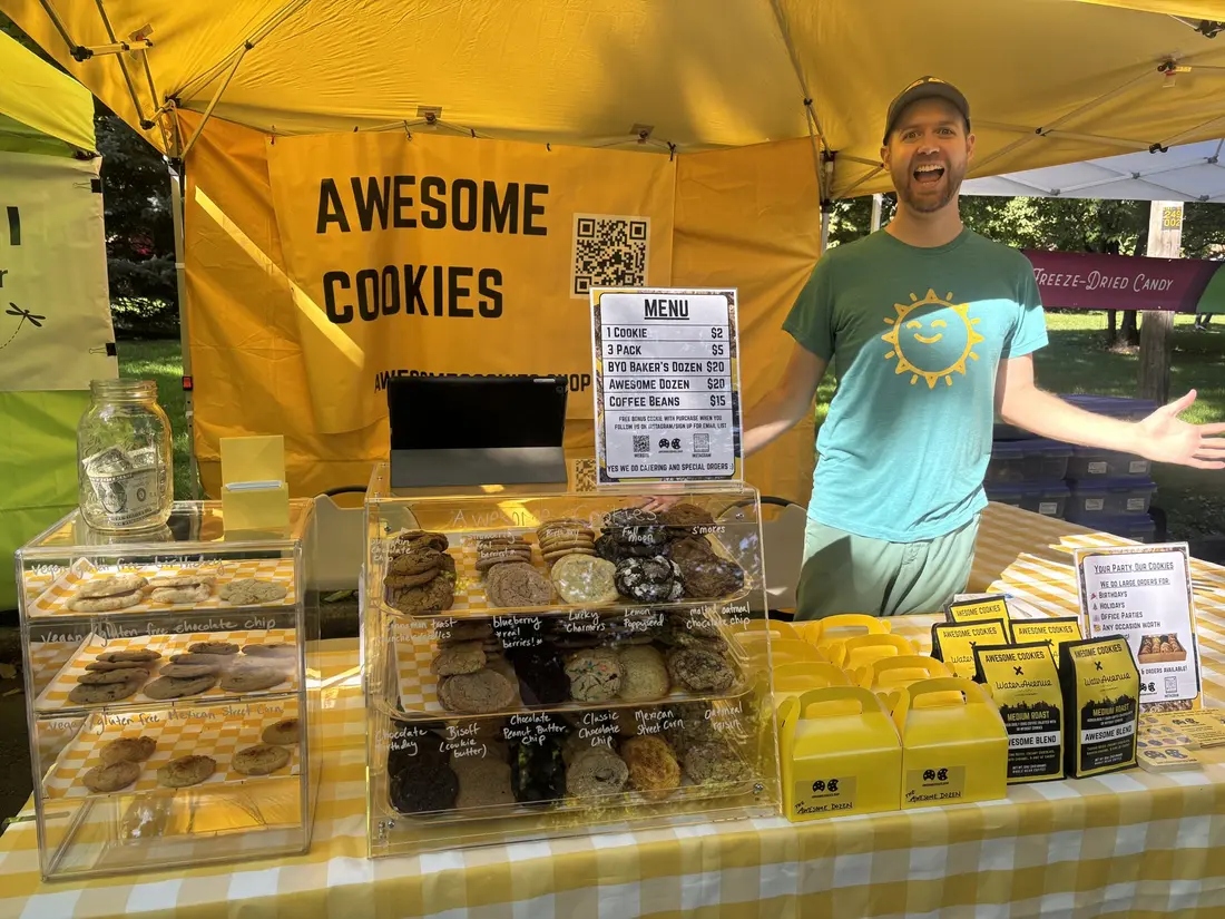 The maker grinning under a yellow tent with trays of cookies and an Awesome Cookies banner.