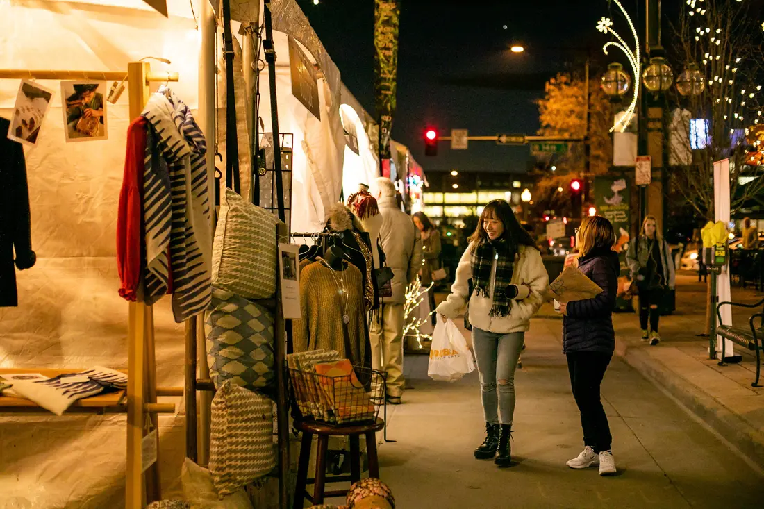 Shoppers at a maker’s tent on a December night, string lights and a lit tree behind.