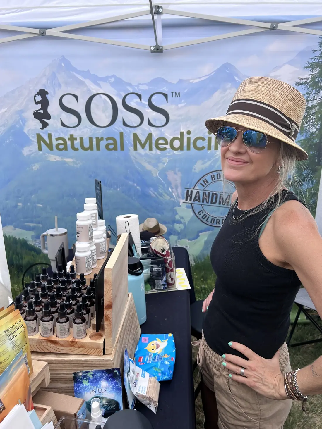 The maker in a straw hat behind her table of bottles under a mountain banner.