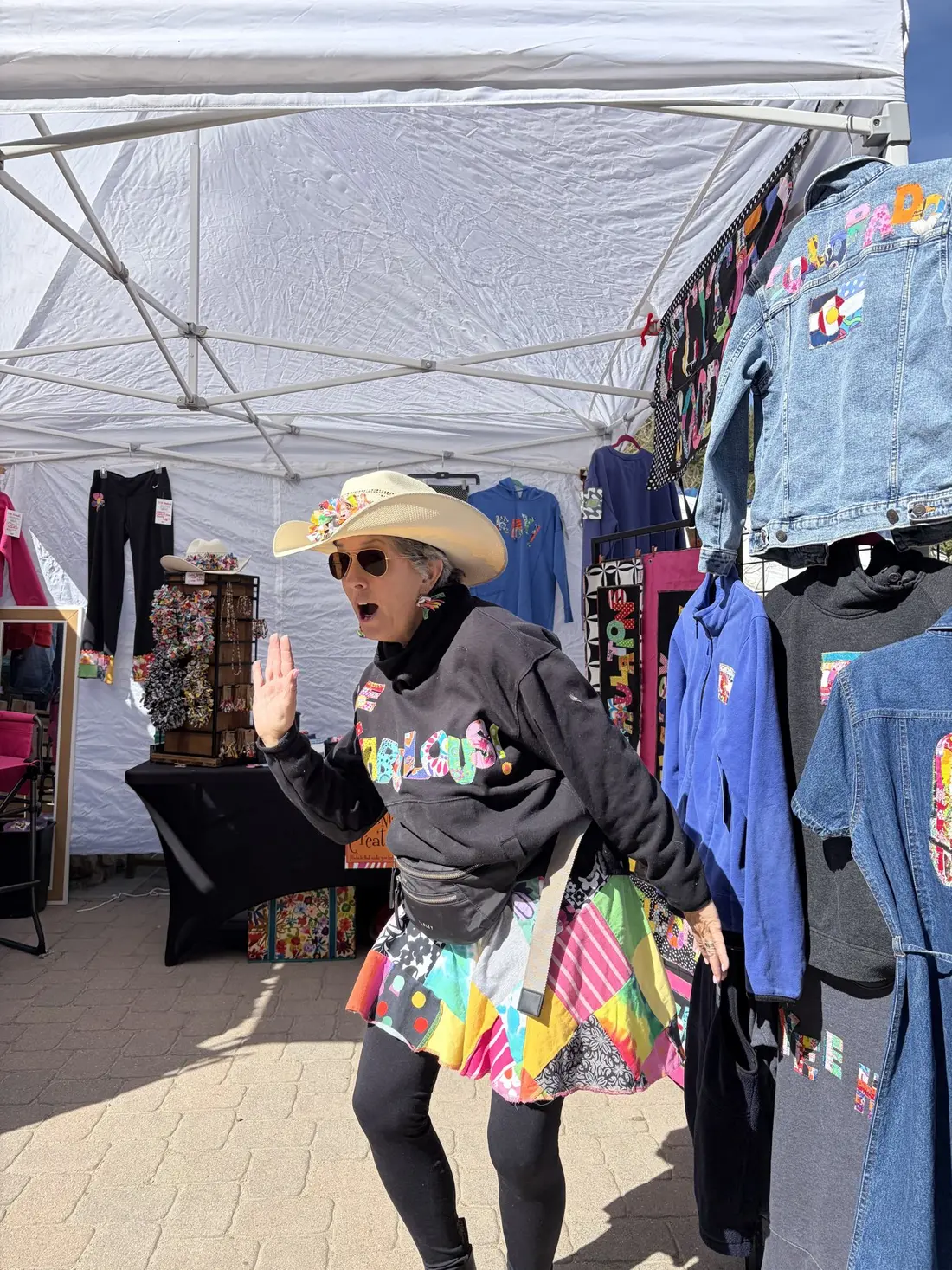 The maker in a cowboy hat and a patchwork skirt, waving from her booth of upcycled clothing.