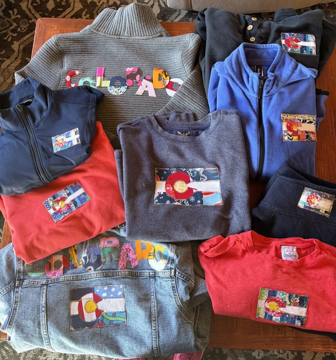 A pile of sweatshirts and jackets, each with a Colorado flag patch on the chest.