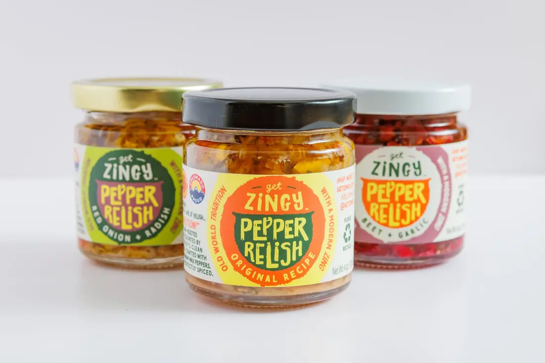 Three jars of Get Zingy pepper relish, red, yellow and green lids, on a white table.