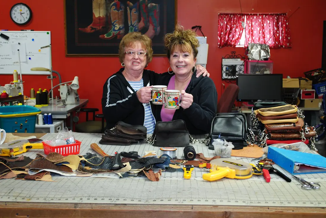Two makers at a workbench covered in leather pieces and tools, holding up a finished bag.