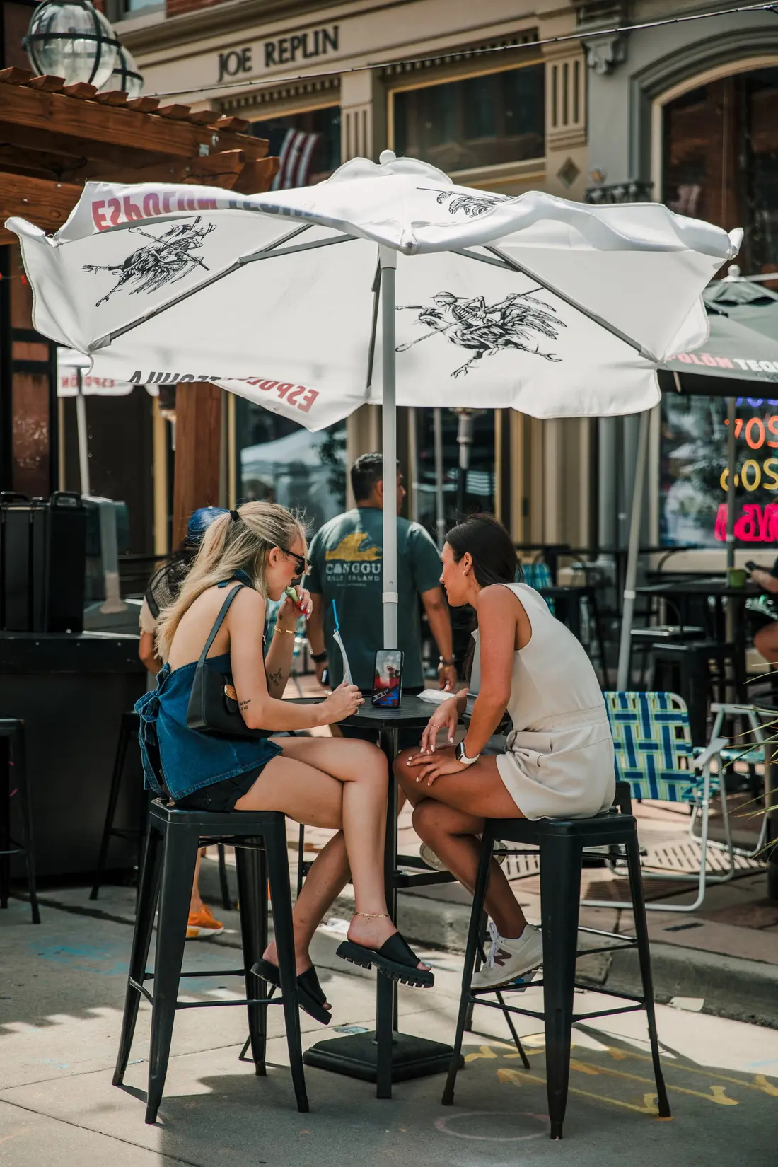 Two friends at a patio table under an umbrella on Larimer Square while the market runs behind them.