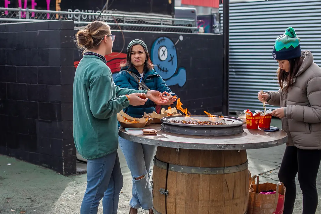 Two friends roasting marshmallows over a fire barrel at a warming station, in daylight.