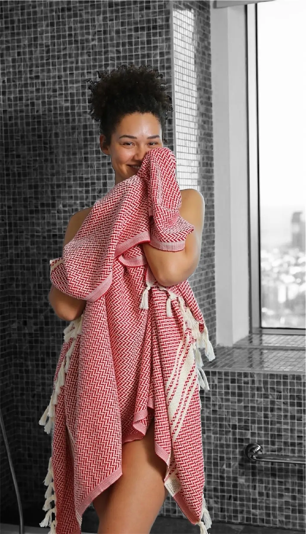 A woman holding a red Turkish towel up to her face in a tiled bathroom.