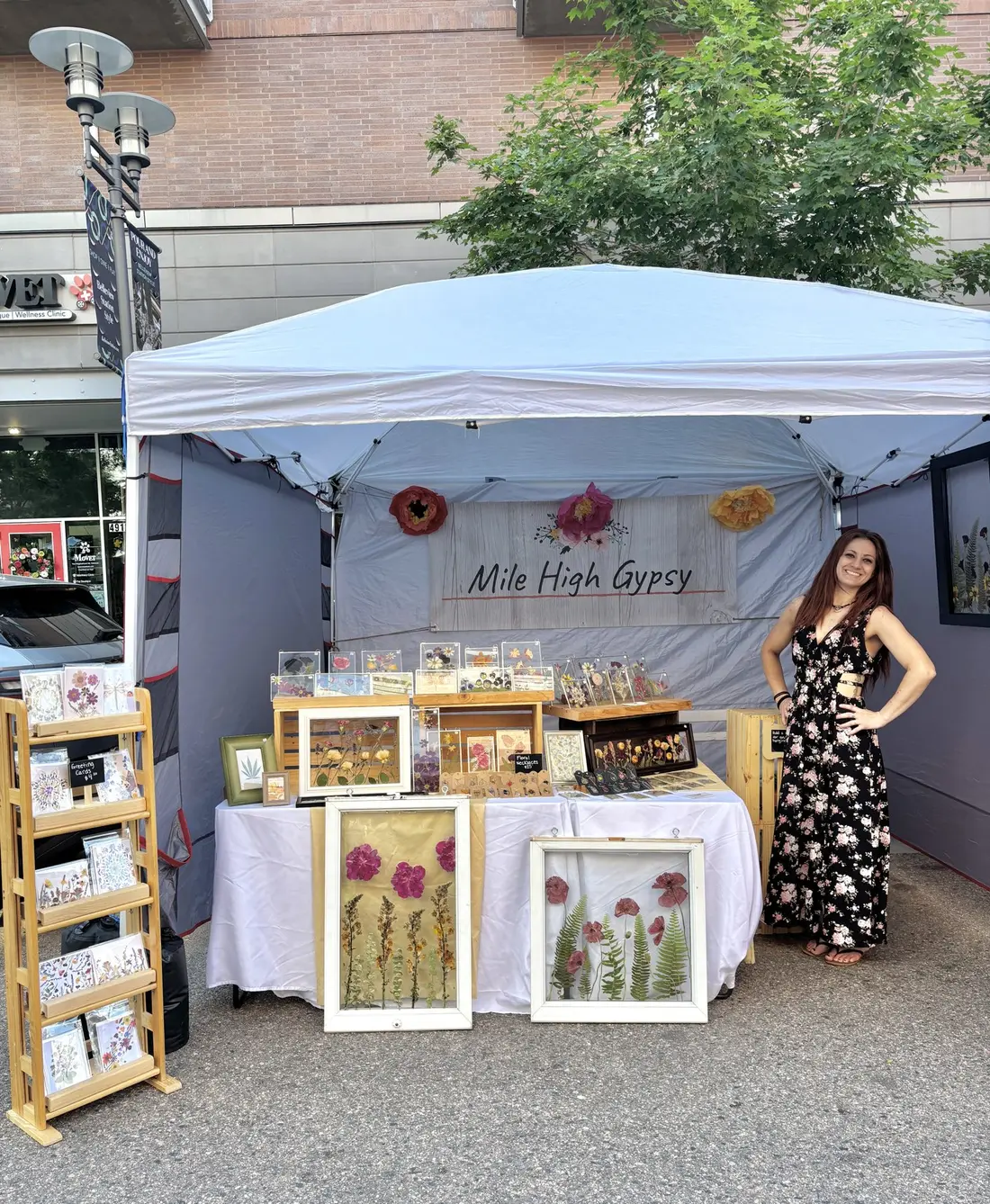 The maker beside her white tent, framed pressed-flower pieces on the table and shelves.