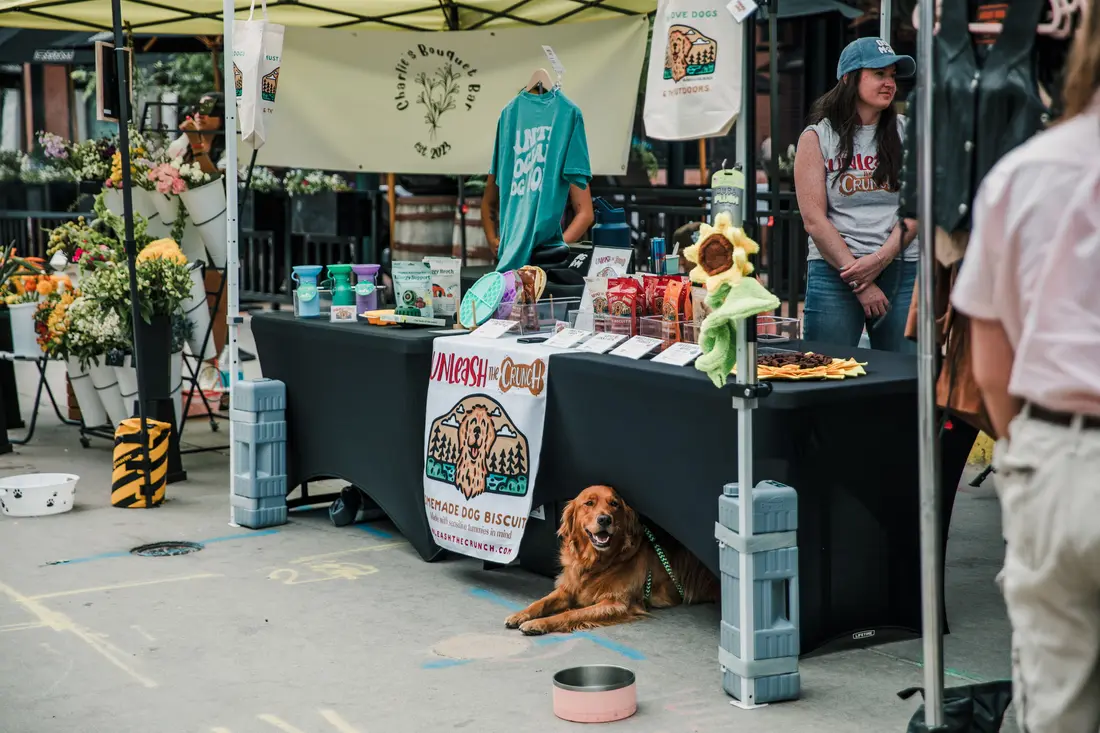 A dog lying under a maker’s table at Larimer Square, its person shopping above.