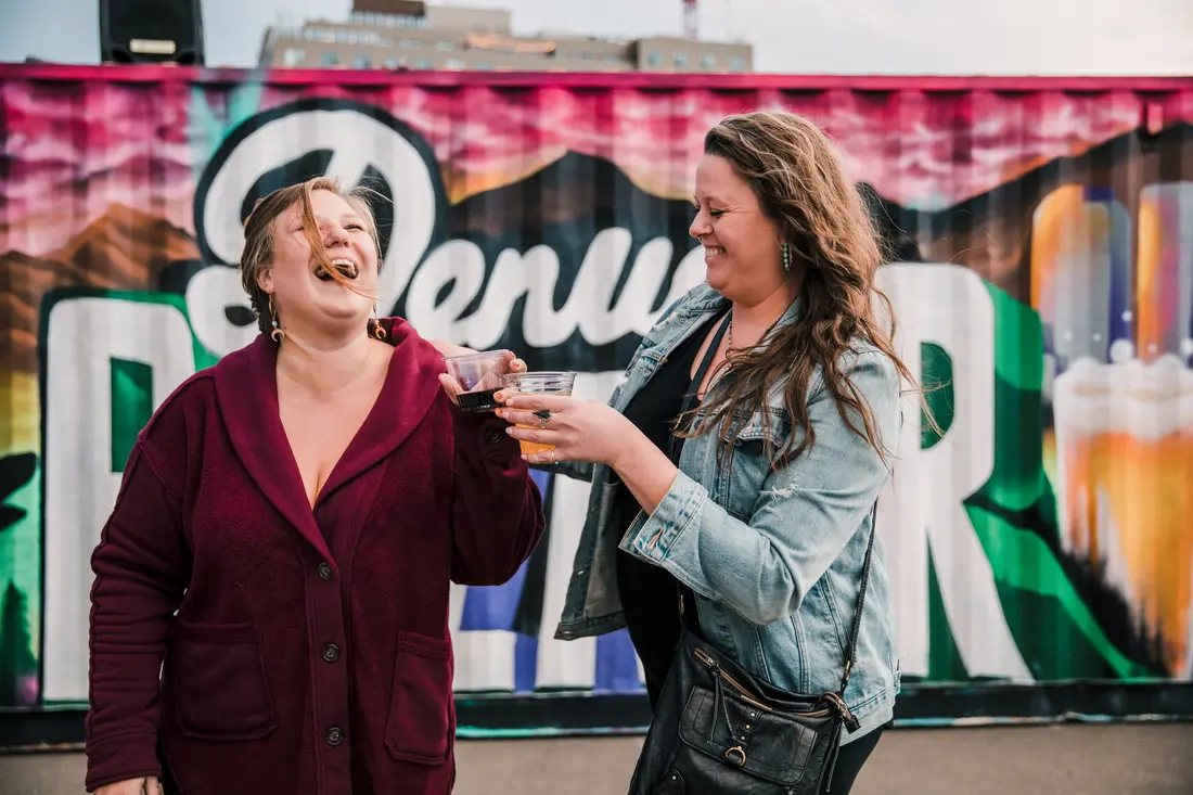 Two women laughing with drinks in hand in front of the Denver BAZAAR banner at the RiNo Holiday BAZAAR.