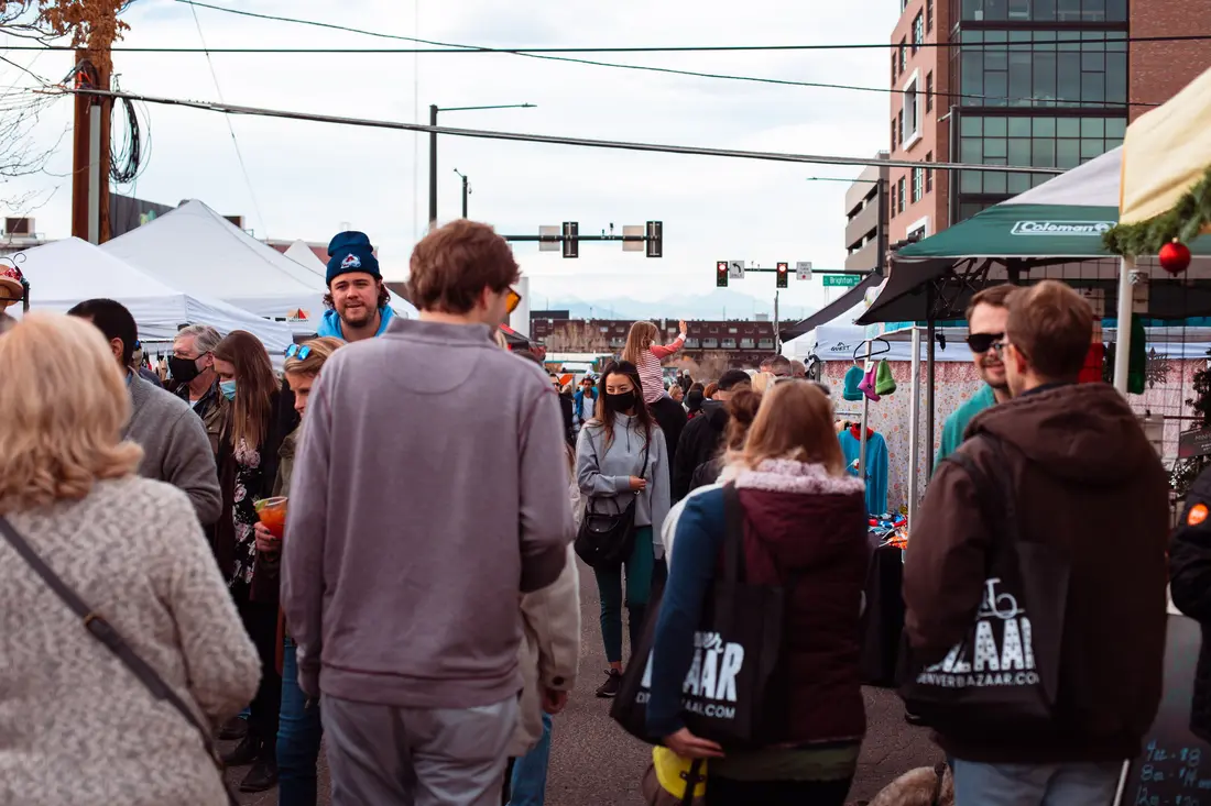 Shoppers between white tents at the RiNo Holiday BAZAAR, the Denver skyline behind.