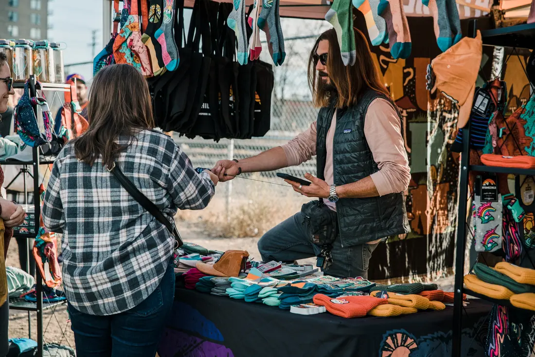 A maker handing a wrapped purchase across the table to a shopper at the RiNo Holiday BAZAAR.