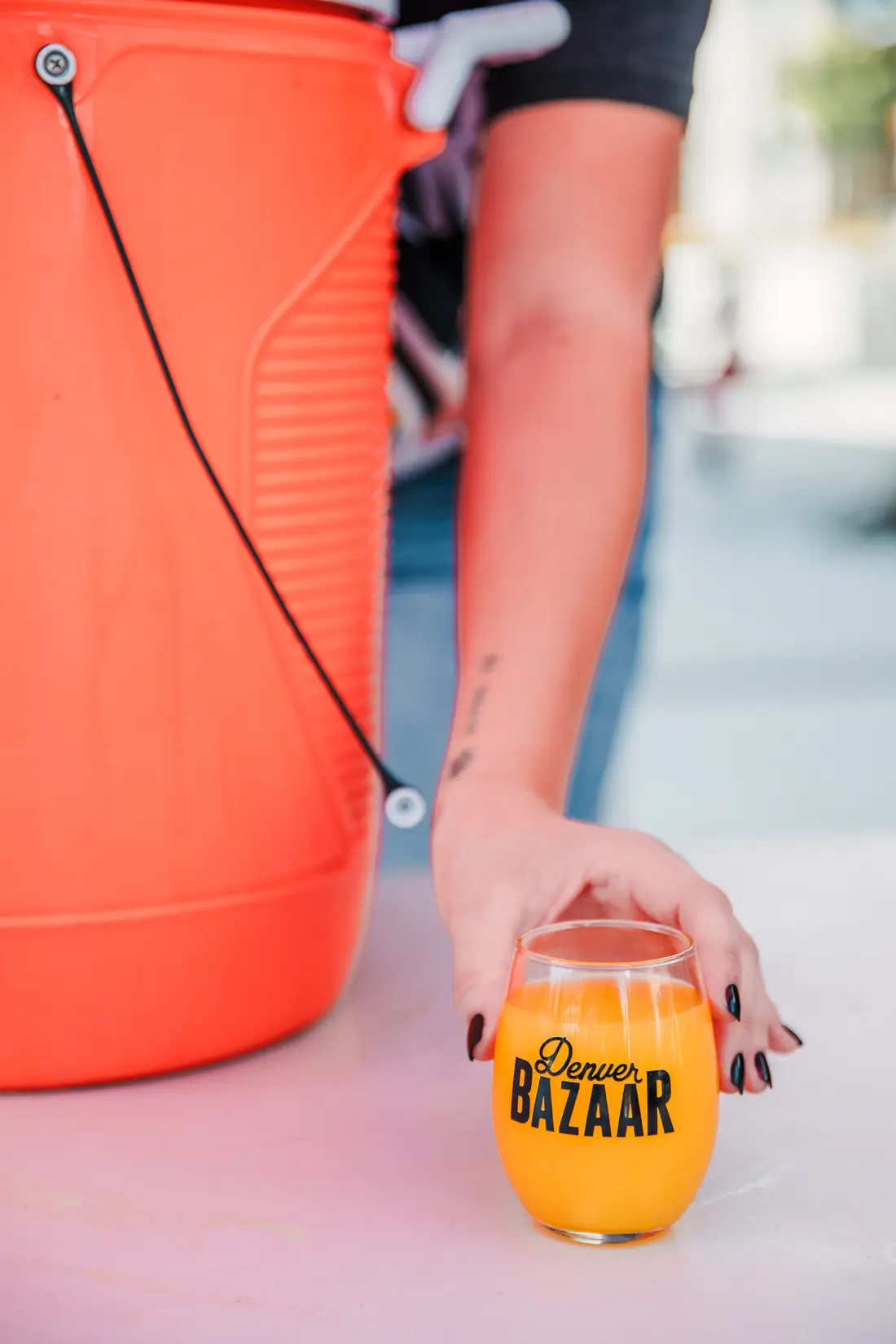 A hand setting a Denver BAZAAR glass of something orange down on an orange cooler at Sloan’s Lake.