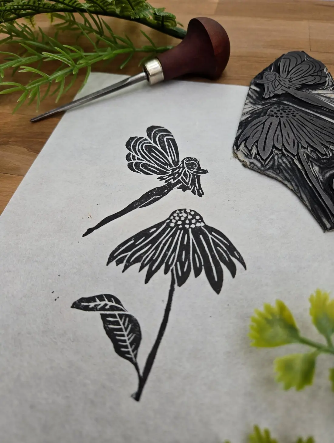 A carved block and a fresh print of a coneflower on a wooden table with the carving tool.