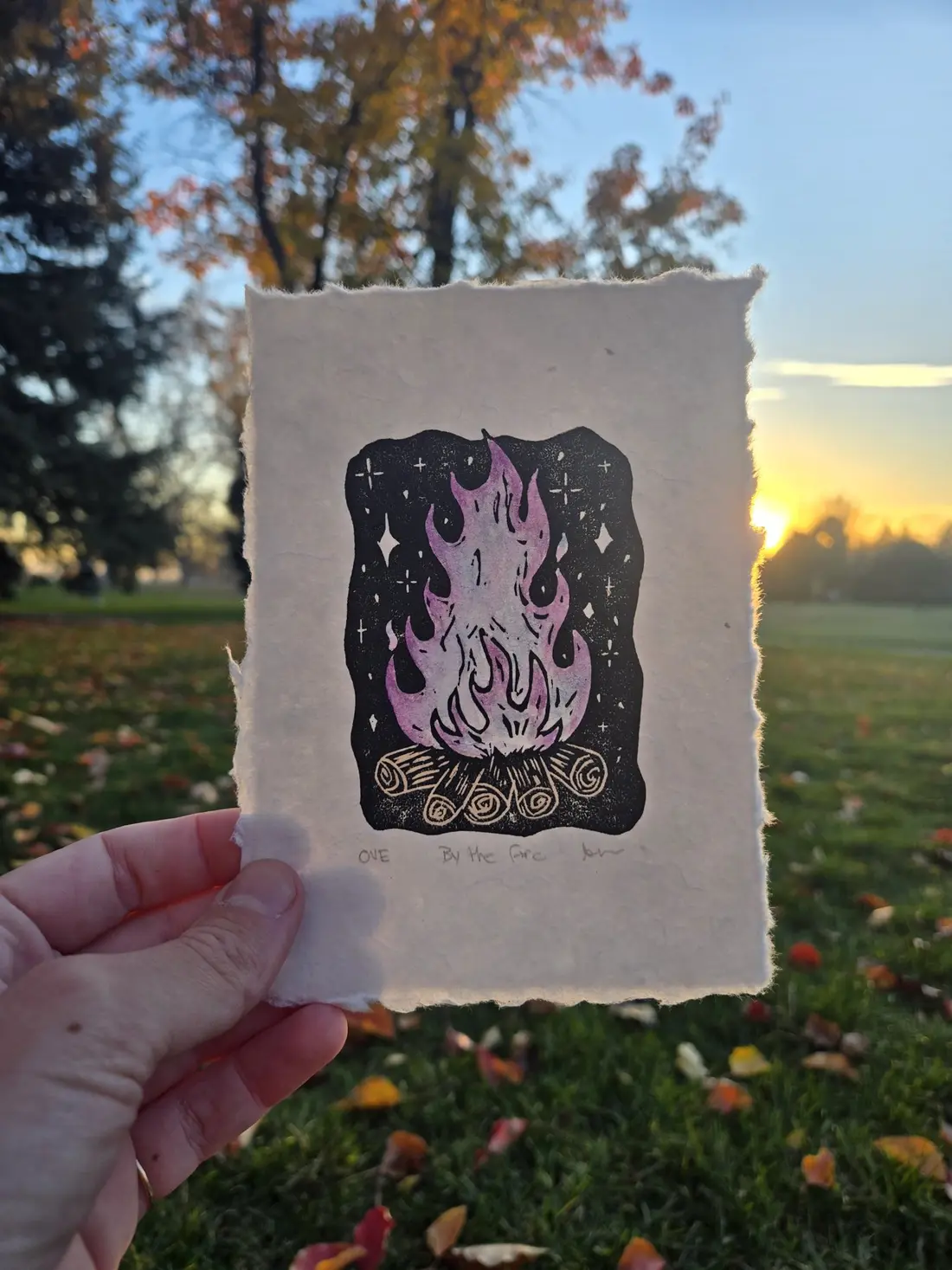 A hand holding up a small print of a cat inside a flame against a sunset in a park.