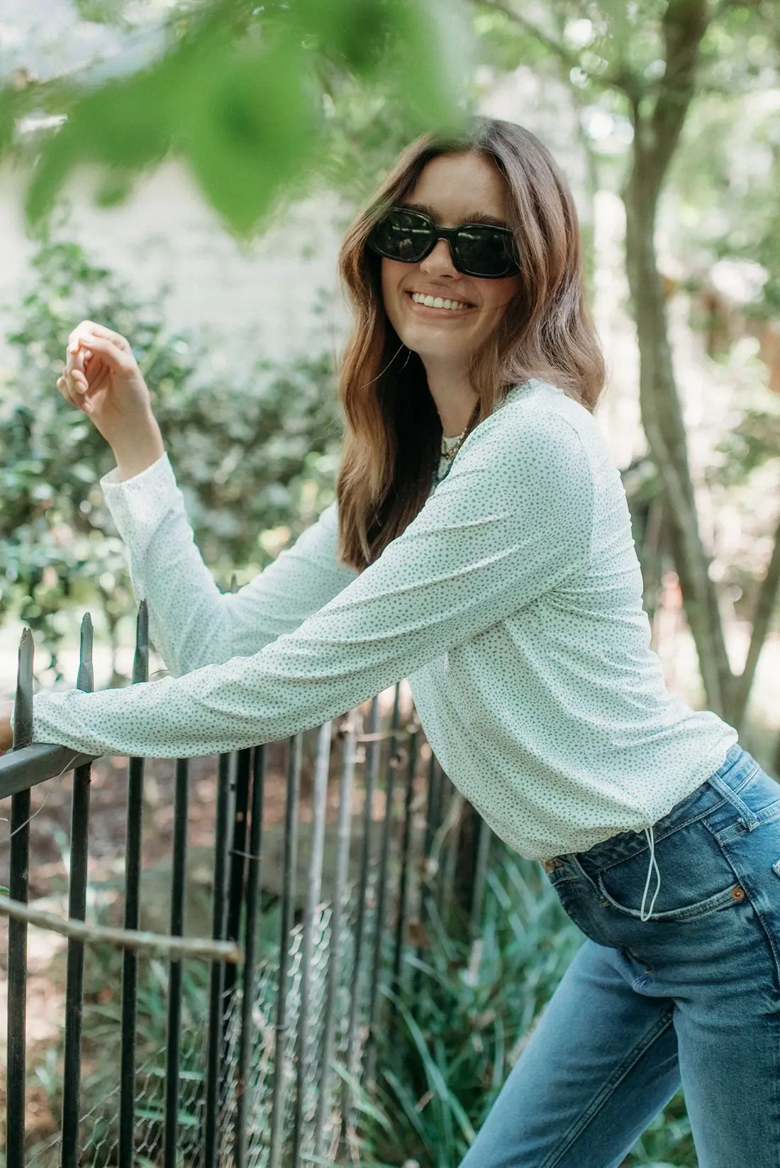 A woman in sunglasses and a white long-sleeve crop shirt leaning on a railing.