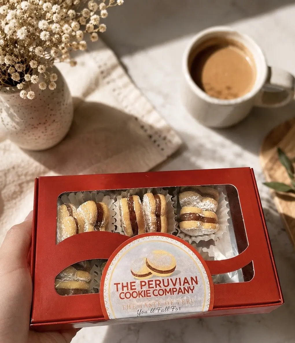 A hand holding a red box of alfajores with The Peruvian Cookie Company label.