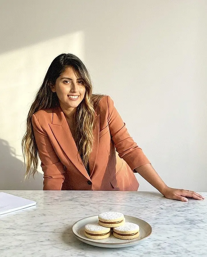 The maker in a rust blazer leaning on a marble counter beside a plate of alfajores.